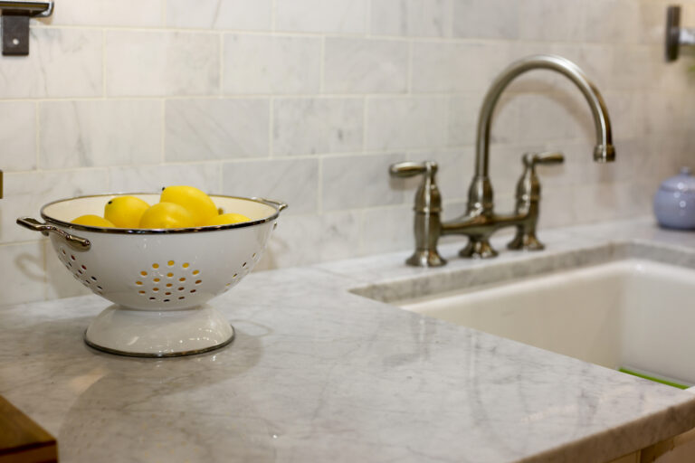 White Carrara marble countertop and backsplash with farmhouse sink, bridge faucet and colander of lemons